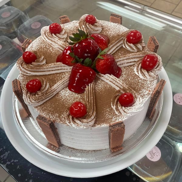 White frosted cake topped with strawberries, cherries, chocolate wafers, and cocoa powder in a display case.