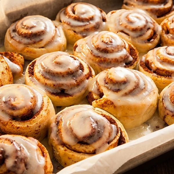 Freshly baked cinnamon rolls with white icing in a baking pan lined with parchment paper.